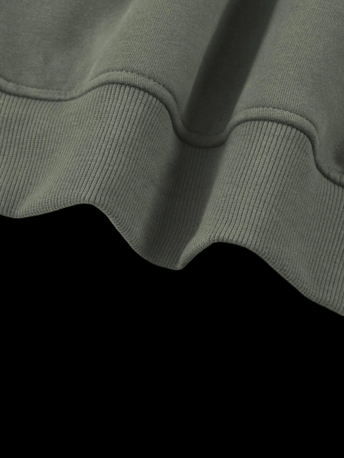 Close-up of a ribbed knit garment, likely a sweatshirt or hoodie, showing the texture and hem detail in a muted olive green color against a plain white background.