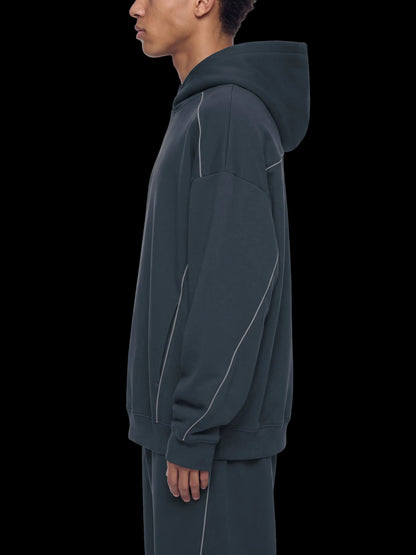 A side-profile view of a person wearing an oversized dark charcoal hoodie with contrasting light piping details, paired with matching dark pants, against a plain white background.
